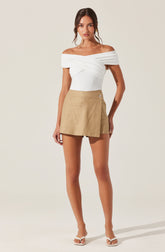 A woman is wearing an off-the-shoulder white top with draped, twisted fabric across the chest, paired with a beige wrap-style mini skirt that has a side button detail. She is also wearing white heeled sandals.